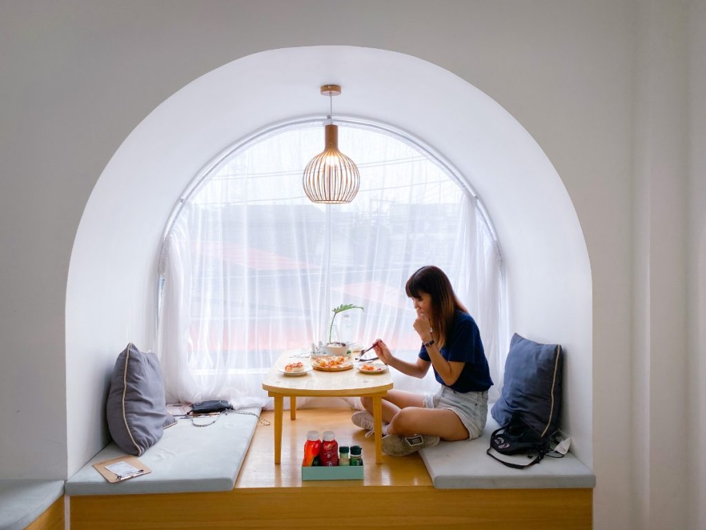 Une femme assise dans sa chambre derrière une fenêtre demi-lune avec du rideau en voile